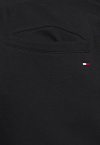 Schwarzer Stoff mit einer flachen Taschenoptik und einem kleinen rot, weiß und blauen gestickten Logo in der unteren Ecke. Glatte Textur.