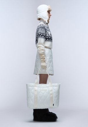 Bolsa de tote blanca acolchada con tirantes con logo, llevada por una figura que viste un suéter de punto y un gorro acolchado a juego. Botas negras de pelaje completan el conjunto.