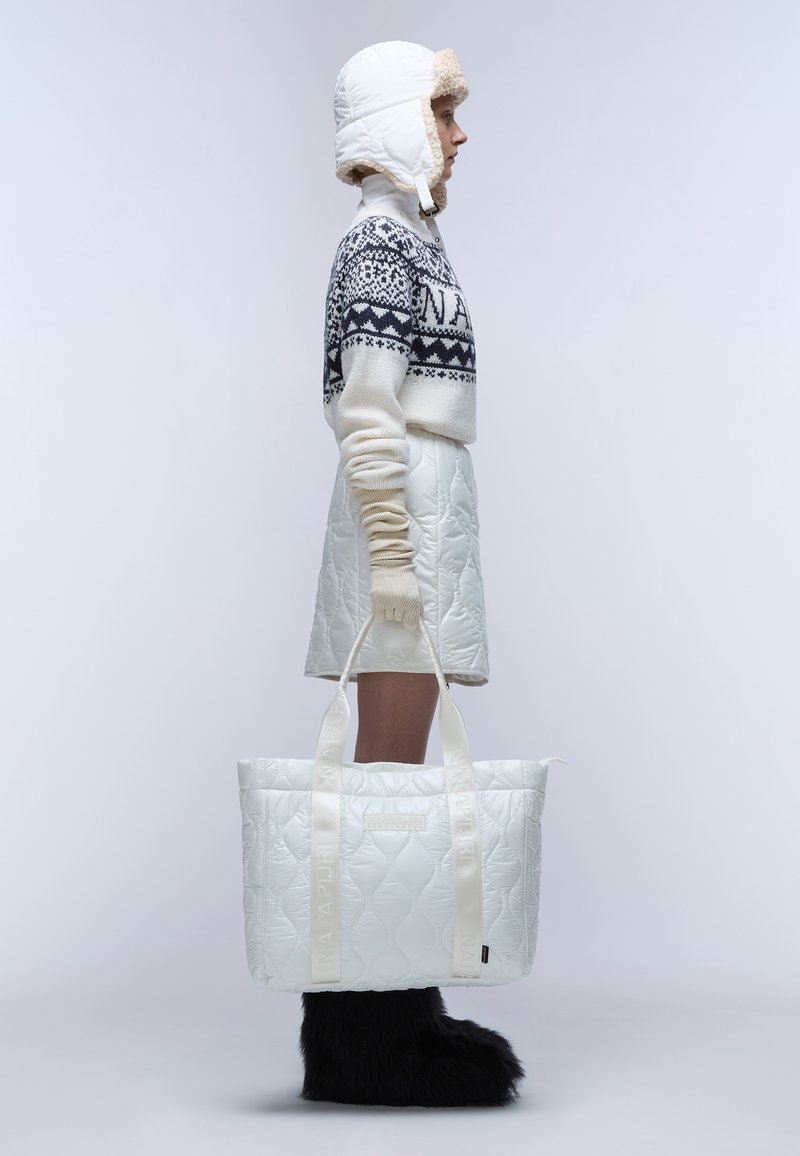 Borsa tote bianca trapuntata con manici con logo, portata da una figura che indossa un maglione lavorato a maglia e un berretto imbottito abbinato. Stivali neri in pelliccia completano il look.