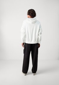 Λευκή φούτερ μπλούζα με κουκούλα και χαλαρή εφαρμογή, συνδυασμένη με μαύρο φαρδύ παντελόνι cargo που διαθέτει πλαϊνές τσέπες και λεπτομέρειες με κορδόνι.