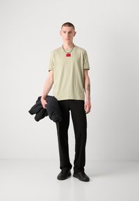 Tricou scurt cu mânecă scurtă de culoare verde deschis, cu un etichet roșu, pantaloni negri, ținând o geacă puf neagră. Sneakerși negri completează ținuta.