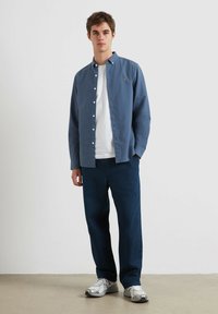 Blauwe lange mouwen shirt met knopen, gedragen over een wit T-shirt, gecombineerd met donkerblauwe broek en zilveren sportschoenen.