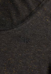 Camisa gris oscuro con superficie texturizada y motas doradas dispersas. Incluye un logotipo bordado en negro en la parte inferior izquierda.