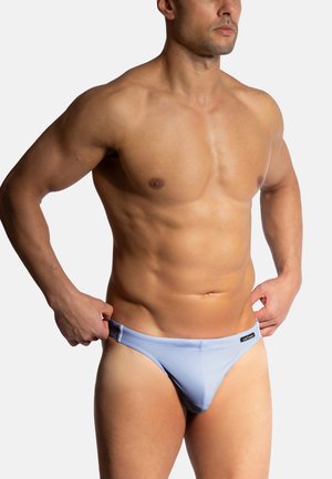 Light blue swim briefs made of smooth fabric, featuring a low-rise cut and a black logo tag on the waistband. Model poses with one hand adjusting.