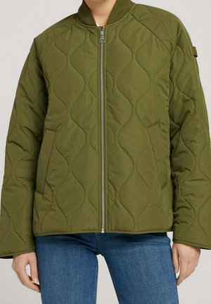 Olivově zelená prošívaná bunda se vlnitým prošitím, předním zipem a bočními kapsami, nošená s modrými džínami na stojící osobě.