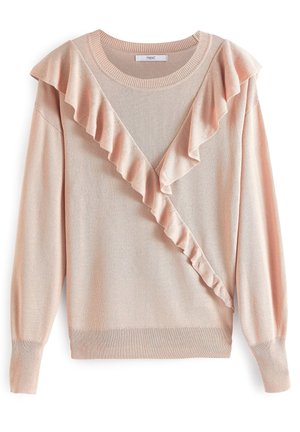 Femme portant un pull rose à volants, un pantalon slim bordeaux, des sandales à talons imprimé serpent et des bracelets, appuyée contre un mur rose clair.