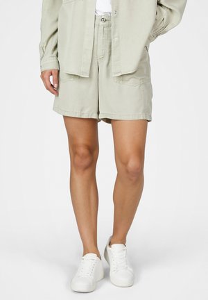 Person trägt beige Shorts mit hohem Bund und großen Fronttaschen, ein passendes beiges Hemd und weiße Sneaker auf einfarbigem Hintergrund.