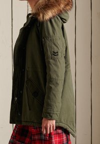 Donna che indossa un parka verde con cappuccio foderato in pelliccia e una gonna a quadri rossi, in piedi di lato contro un muro di legno bianco.
