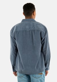 Camisa de manga larga azul, tela texturizada, cuello abotonado, detalle en el yugo trasero y dobladillo recto, combinada con jeans azul claro.