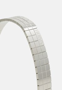 Bracelet en métal argenté avec un motif en grille, présentant une finition lisse. Logo "HUGO" gravé visible sur le côté.