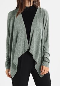 Světle zelený cardigan s volně draperovaným předním dílem, dlouhými rukávy a měkkou texturou. Nositý přes černý rolák, se volným střihem.
