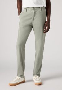 Pantaloni verde chiaro con vestibilità slim, realizzati in tessuto testurizzato, dotati di tasche frontali e chiusura con bottone. Abbinati a scarpe da ginnastica bianche.