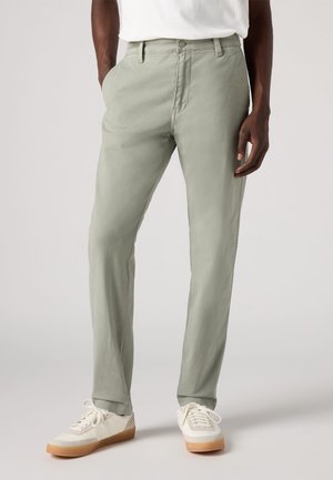 Pantaloni verde chiaro con vestibilità slim, realizzati in tessuto testurizzato, dotati di tasche frontali e chiusura con bottone. Abbinati a scarpe da ginnastica bianche.