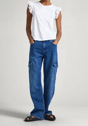 Henkilö, joka pukeutunut valkoiseen liehulahkeiseen toppiin, leveälahkeisiin sinisiin farkkuratsastushousuihin, joissa on sivutaskut, ja mustiin avojalkakenkiin, seisoo yksiväristä taustaa vasten.