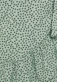 Tecido verde com um pequeno padrão floral preto. Apresenta uma bainha com babados. Textura macia com uma aparência leve.