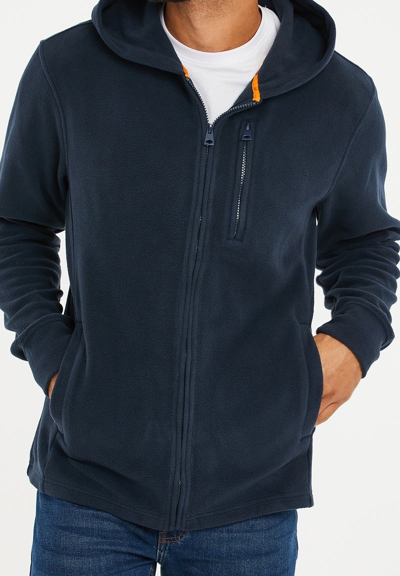 Homme portant un sweat à capuche zippé en polaire bleu marine avec les mains dans les poches et un t-shirt blanc en dessous, montré du cou aux hanches.