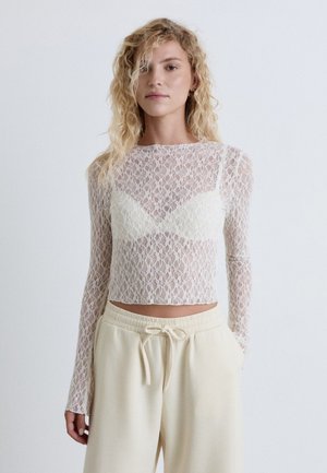 Jeune femme aux cheveux blonds bouclés portant un haut blanc en dentelle à manches longues par-dessus un soutien-gorge blanc et un pantalon crème avec cordon de serrage.