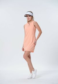Perzikkleurige jurk met korte mouwen, een ritskraag, getextureerde zijpanelen en witte sneakers. Het model draagt een lichte zonnebril.