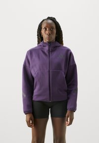 Lila zip-up jacka med hög krage, med en frontficka och grå accentränder på ärmarna. Mjuk tygtextur.