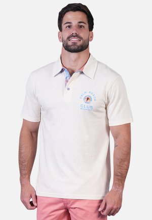 Homme portant un polo crème avec le logo "Palm Beach Club" et un short rose, debout sur un fond uni.