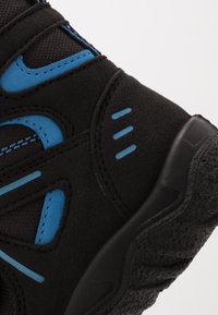 Svart träningsskor med blå accenter, med en texturerad överdel och förstärkt tå. Designen inkluderar eleganta linjer och kontrasterande sömmar.