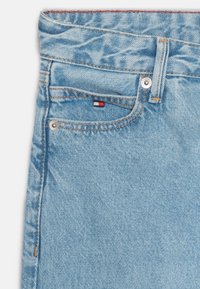 Vaqueros de mezclilla azul claro con un botón plateado visible, costuras naranjas y una pequeña etiqueta con el logo en rojo, blanco y azul en el bolsillo delantero.