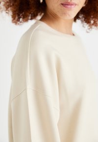 Beige Sweatshirt mit lockerer Passform. Merkmale einen runden Ausschnitt, überschnittene Schultern und eine weiche Textur. Minimale Nähte.