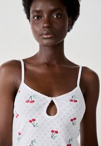 Camisola bianca con scollatura a chiave, bordi in pizzo e un motivo a ciliegie con foglie verdi su un tessuto testurizzato.