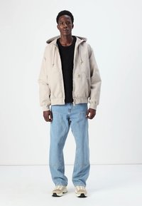 Beige hoodie met ritssluiting, voorkantzakken en een gestructureerde zwart gebreide trui. Lichtblauwe jeans met gestikte patronen, witte sneakers.