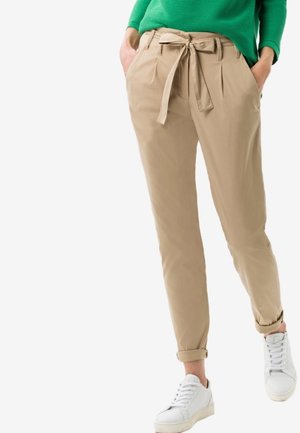 Mujer con pantalones beige de cintura fruncida con cinturón anudado, puños doblados, suéter verde y zapatillas blancas, con las manos en los bolsillos, caminando hacia adelante.