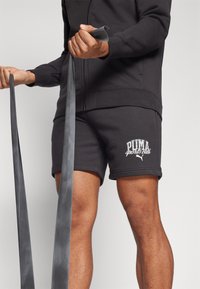 Čierna bavlnená mikina na zips a zodpovedajúce šortky s bielym logom "PUMA sports club". Nositeľ drží čierne cvičebné gumy.
