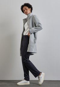 Grå ullkappa med hög krage, tryckknappar och sidfickor, bärs över en vit topp och mörka texturerade byxor, kombinerad med vita sneakers.