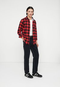 Camicia flanella a quadri rossi e neri, t-shirt bianca sotto, abbinata a jeans scuri e sneakers nere con strisce bianche.