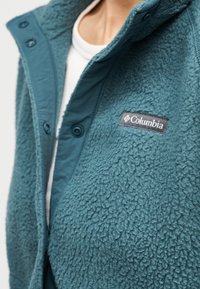 Κοντινό πλάνο ατόμου που φοράει γαλάζιο φλις μπουφάν με το λογότυπο της Columbia πάνω από λευκό πουκάμισο, εμφανίζοντας την υφή και το μερικώς κλειστό εμπρόσθιο μέρος.