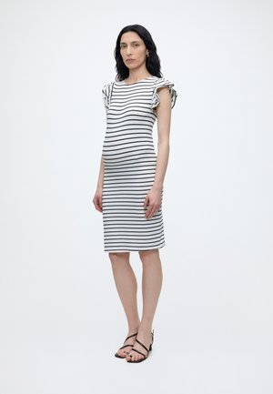 Γυναίκα στέκεται φορώντας ένα λευκό φόρεμα μέχρι το γόνατο με μαύρες οριζόντιες ρίγες και βολάν στα κοντά μανίκια, συνδυασμένο με μαύρα σανδάλια με λουράκια.