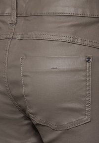 Tajta beige byxor med en slät textur, featuring en bakficka, framträdande sömmar och en glänsande metallknapp accent.