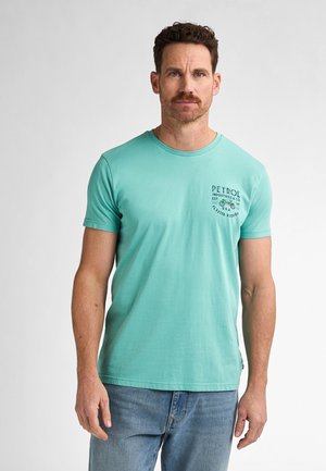 Camiseta de algodón de manga corta en turquesa claro, con cuello redondo y pequeño logo impreso en el lado izquierdo del pecho. Combinada con vaqueros azules.
