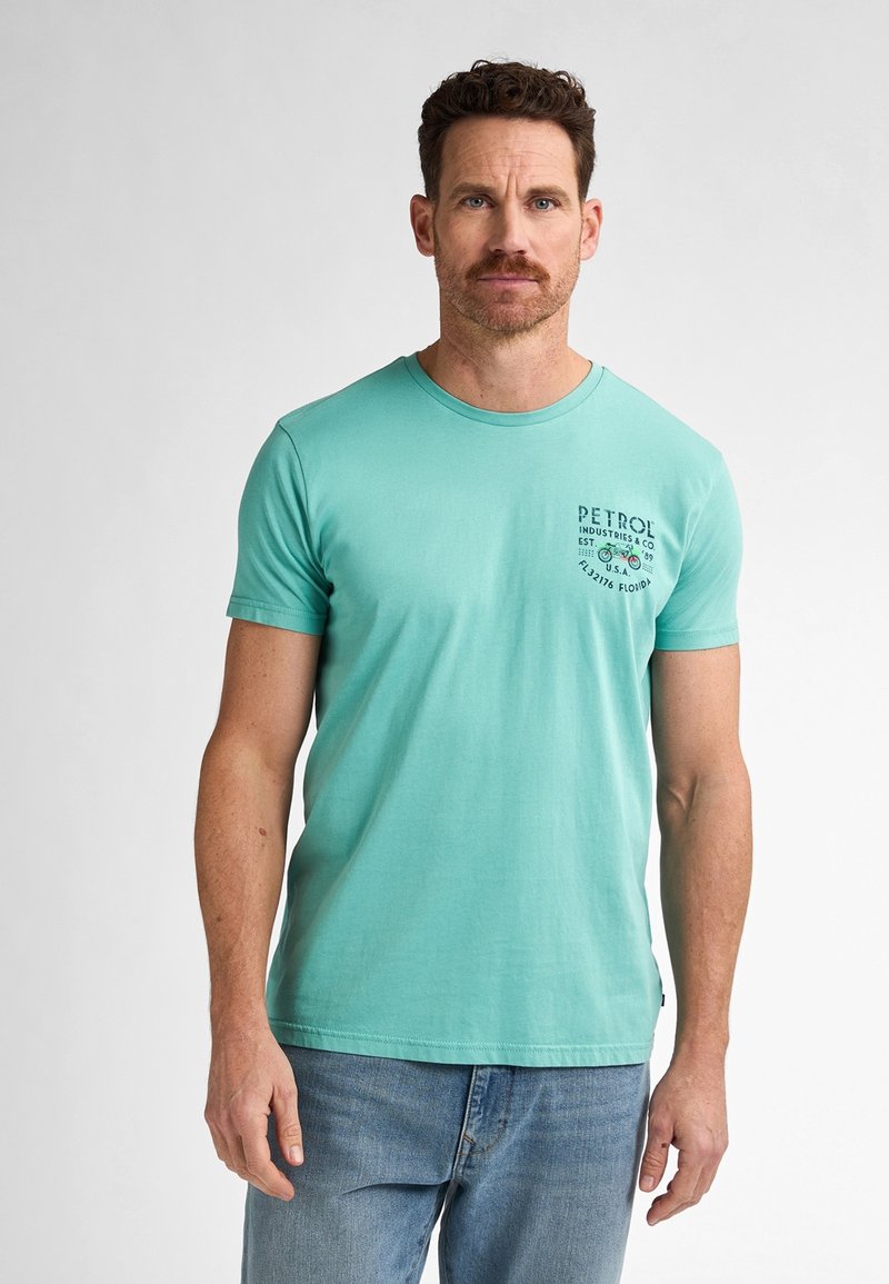 Světle tyrkysové tričko s krátkým rukávem z bavlny, s kulatým výstřihem a malým tištěným logem na levé straně hrudi. Nosené s modrými džíny.