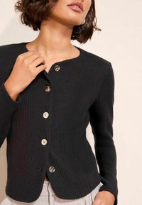 Crna rebrasta kardigan s okruglim izrezom, zatvaranjem na pet dugmadi i suptilnim sjajem na dugmadima. Tkanina ima teksturirani izgled.