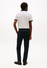 Homme les mains dans les poches, portant un polo blanc à rayures bleu marine et un pantalon bleu marine, debout, de dos, sur un fond clair uni.