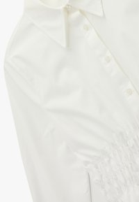 Camisa blanca abotonada con cuello clásico, con detalles fruncidos en la cintura y textura de tela lisa.