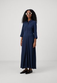 Tumši zils, garām piedurknēm, pogājams kleita no mīksta auduma. Tam ir pieguļošs augšdaļa un plūstoša svārku daļa. Modelis valkā melnas kurpes un baltas saulesbrilles.