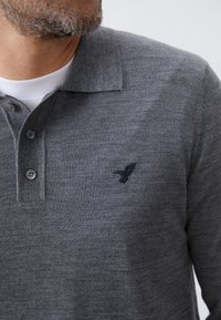 Grå polo t-shirt med strikket tekstur, to-knaps lukning og et broderet fuglelogo i mørk grå på venstre bryst.