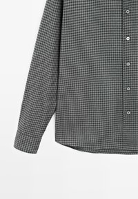 Grå och svart rutig långärmad skjorta med knappslå fram, platt krage och rundad nederkant. Tillverkad av ett mjukt tyg med texturerat mönster.