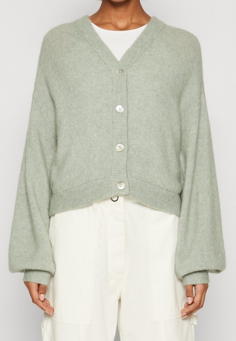 Světlé zelené krátké cardigan z měkké, texturované látky. Má výstřih do V, dlouhé balónové rukávy a na přední straně pět průhledných knoflíků.
