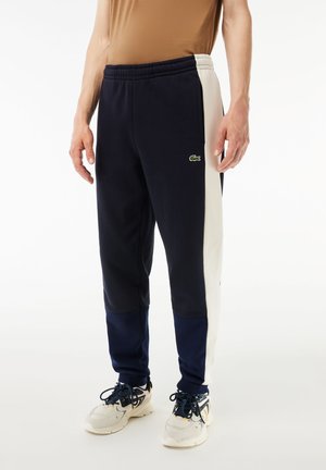 Pantalones de chándal azul marino con paneles laterales blancos, cinturilla elástica y pequeño logo verde. Tejido suave con un diseño relajado.