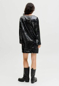 Crna haljina od šljokica s dugim rukavima, s okruglim izrezom i slobodnim krojem, uparena s crnim kožnim čizmama do koljena.