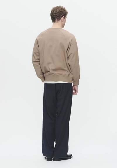 Felpa marrone con collo a giro e maniche raglan, indossata sopra una camicia bianca, abbinata a pantaloni larghi blu navy e scarpe nere, vista da dietro.