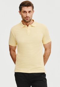 Żółta koszulka polo z krótkim rękawem i kołnierzykiem, wykonana z bawełny. Posiada subtelne logo po lewej stronie klatki piersiowej. Połączona z czarnymi spodniami.
