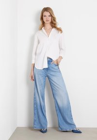 Camisa blanca de botones de manga larga combinada con unos vaqueros de tiro alto y pierna ancha de color azul claro, y zapatos azules de punta con acentos decorativos.
